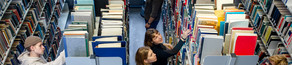 Students stand next to several bookshelves in the library and searching for books.